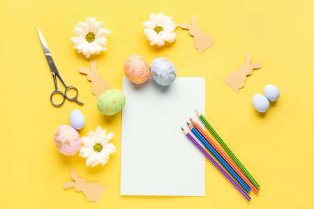 Blank paper sheet with pencils, Easter eggs, rabbits and daisy flowers on yellow backgroundの写真素材