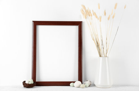Blank frame, vase with dried flowers and Easter eggs on table near white wallの写真素材