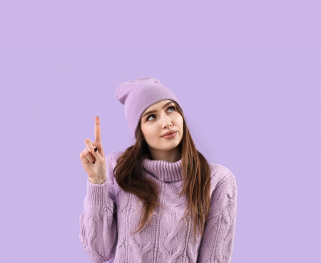 Pretty young woman in knitted sweater and hat pointing at something on lilac backgroundの写真素材