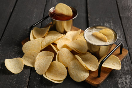 Board with delicious potato chips and sauces on black wooden background