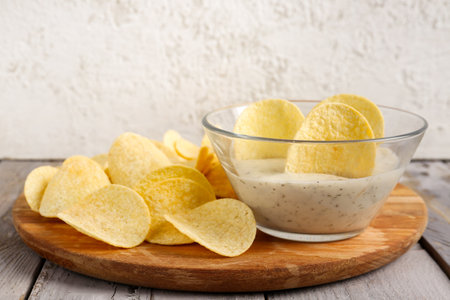 Board with delicious potato chips and bowl of sauce on wooden table against gray backgroundの写真素材