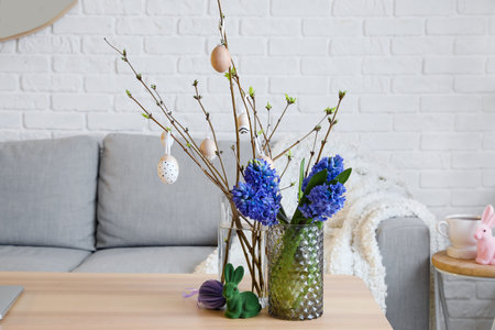 Vases with flowers, tree branches, Easter eggs and rabbits on table in living room