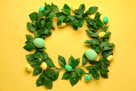 Frame made of green plant branches and painted Easter eggs on yellow backgroundの写真素材