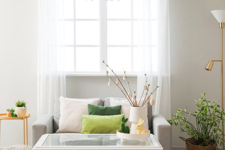 Vase with tree branches, Easter eggs and rabbits on table in light living roomの写真素材