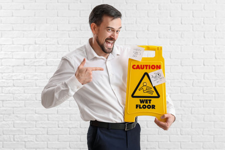 Young man pointing at caution sign with sticky papers on white brick background. April Fools' Day...
