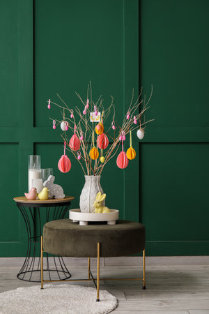 Table, pouf with tree branches and Easter eggs in vase near green wall