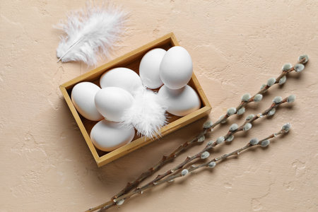 Wooden box with white Easter eggs, feathers and willow branches on color background