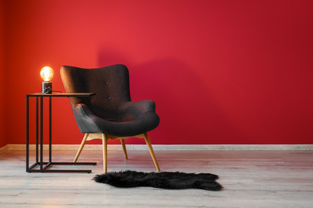 Stylish gray armchair, black fur rug and table with glowing lamp near red wallの写真素材