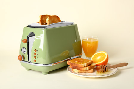 Bread slices in modern toaster, glass of juice, orange and tasty toasts with honey on beige backgroundの写真素材