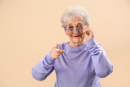 Senior woman in sunglasses pointing at viewer on beige backgroundの写真素材