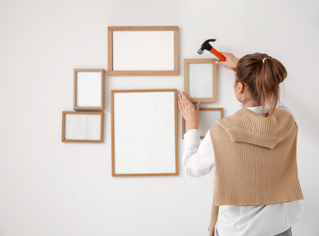 Young woman with hammer hanging blank frame on light wall at home, back viewの写真素材