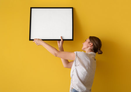 Young woman hanging blank frame on yellow wall at homeの写真素材