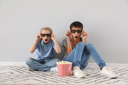 Shocked little boy and his sister in 3D glasses watching cartoons on TV near light wallの写真素材