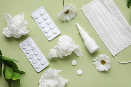 Nasal drops with pills, flowers, medical mask and tissues on green background. Seasonal allergy...