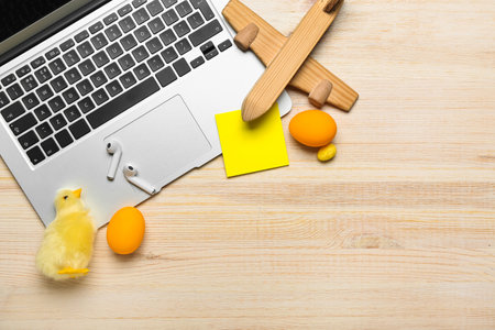 Laptop with earphones, toy airplane, chicken, sticky note and Easter eggs on light wooden background