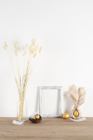 Blank frame, vase with dried flowers and Easter eggs on table near white wallの写真素材