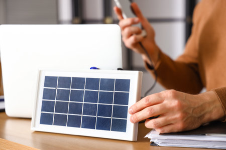 Young man with portable solar panel charging mobile phone in office, closeupの写真素材
