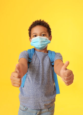 Little African-American schoolboy in medical mask showing thumb-up gesture on color background. Coronavirus epidemicの写真素材
