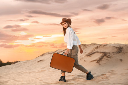 Beautiful fashionable young woman with suitcase in desertの写真素材