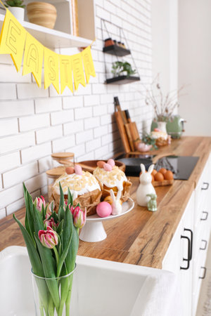 Vase with tulip flowers, Easter cakes and bunnies on kitchen counter near white brick wall