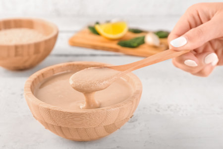 Female hand with spoon and bowl of tasty tahini on light wooden table, closeupの写真素材