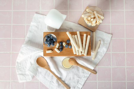 Board with delicious wafer rolls, blueberries and condensed milk on pink tile background