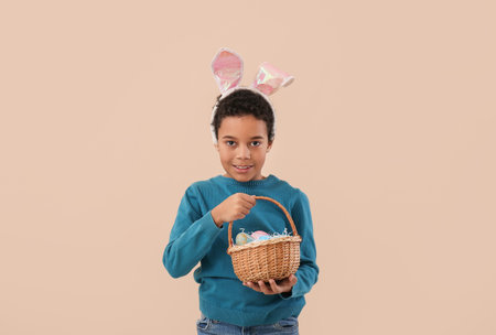 African-American little boy in bunny ears with basket of Easter eggs on beige background