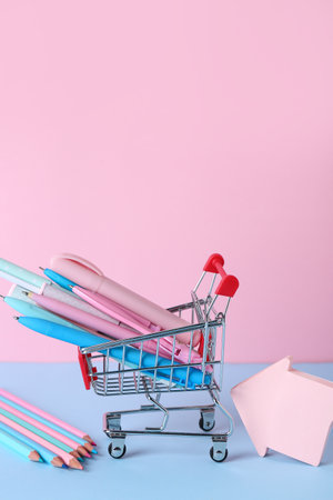 Shopping cart with different pens, pencils and sticky notes on color backgroundの写真素材