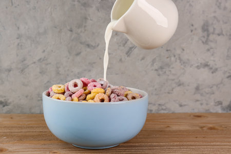 Pouring of milk from pitcher into bowl with colorful cereal rings on wooden table near gray grunge wallの写真素材