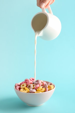 Female hand pouring milk from pitcher into bowl with colorful cereal rings on turquoise backgroundの写真素材