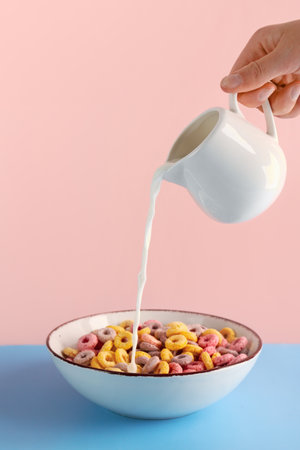 Female hand pouring milk from pitcher into bowl with colorful cereal rings on blue table against pink backgroundの写真素材
