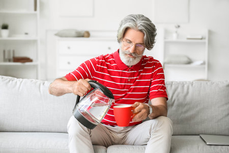Mature man pouring hot water from electric kettle into cup at homeの写真素材