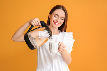Young woman pouring hot boiled water from electric kettle into cup on color backgroundの写真素材