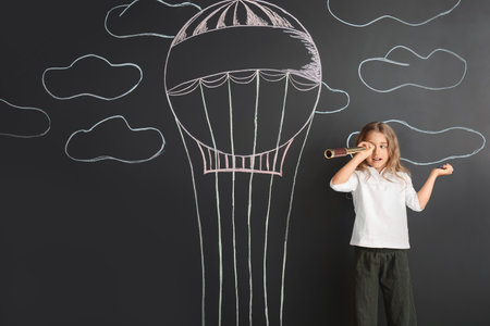 Cute little girl watching in spyglass on black wall with drawn hot air balloonの写真素材