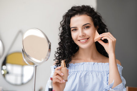 Beautiful young woman applying foundation at homeの写真素材