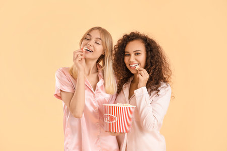 Beautiful young women in pajamas eating popcorn on color backgroundの写真素材