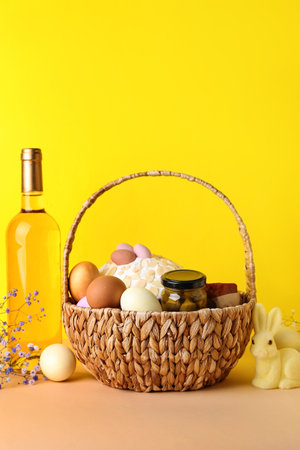 Basket with Easter eggs, cake and bottle of wine on beige table against yellow backgroundの写真素材
