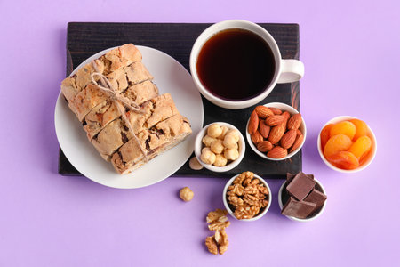 Board with biscotti cookies, chocolate, nuts, dried apricots and cup of coffee on lilac backgroundの写真素材