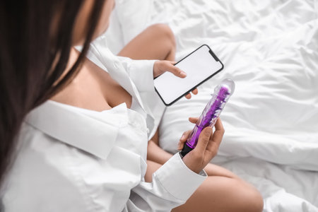 Young woman with vibrator and mobile phone in bedroom, closeupの写真素材