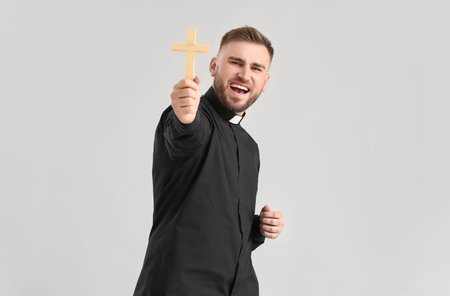 Emotional young priest with cross on light background