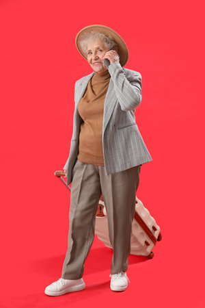 Senior woman with suitcase talking by mobile phone on red backgroundの写真素材