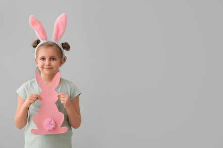 Little girl in bunny ears with paper Easter rabbit on gray backgroundの写真素材