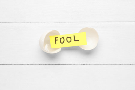 Paper with word FOOL and egg shells on white wooden backgroundの写真素材