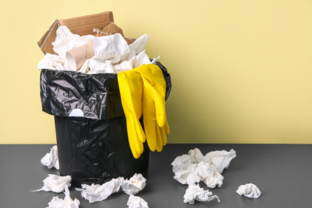Rubbish bin with paper garbage and rubber gloves near yellow wallの写真素材
