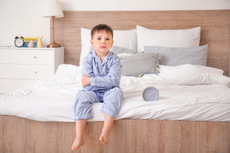 Little boy suffering from sleep disorder with alarm clock in bedroomの写真素材