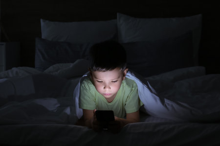Little boy suffering from sleep disorder with mobile phone in bedroomの写真素材