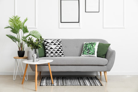 Cozy sofa with cushions and houseplants on tables near white wallの写真素材