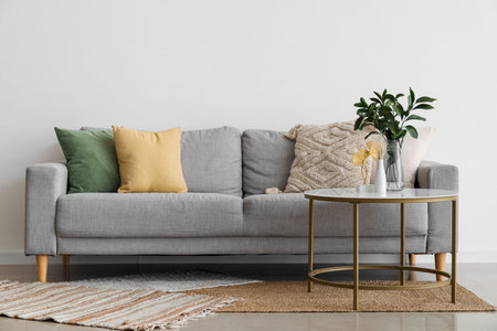 Cozy gray sofa with cushions and plant branches in vase on table near light wall
