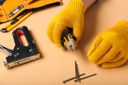 Male hands in gloves holding pliers and nail on beige backgroundの写真素材
