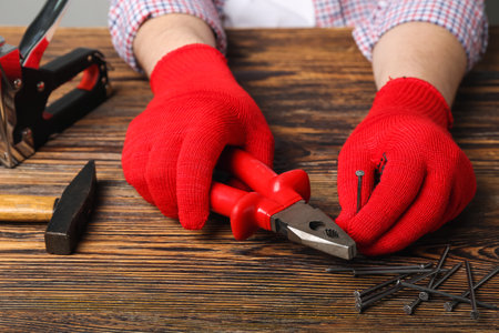 Male hands in gloves holding pliers and nail near wooden tableの写真素材